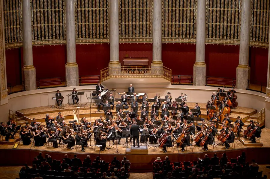 Veduta dall'alto di una sala da concerto storica con colonne, velluti rossi e ori: orchestra sinfonica al completo, direttore al centro e pubblico in platea