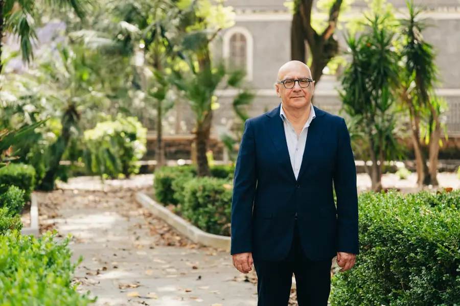 Ritratto orizzontale a figura intera: in piedi al centro di un vialetto di giardino, palme e facciata di un edificio antico sullo sfondo, sguardo in camera