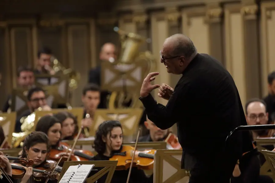 Di tre quarti da dietro, dirige a mani nude sotto una luce calda dorata, davanti a una giovane orchestra di archi e ottoni in una sala dalle decorazioni ornate