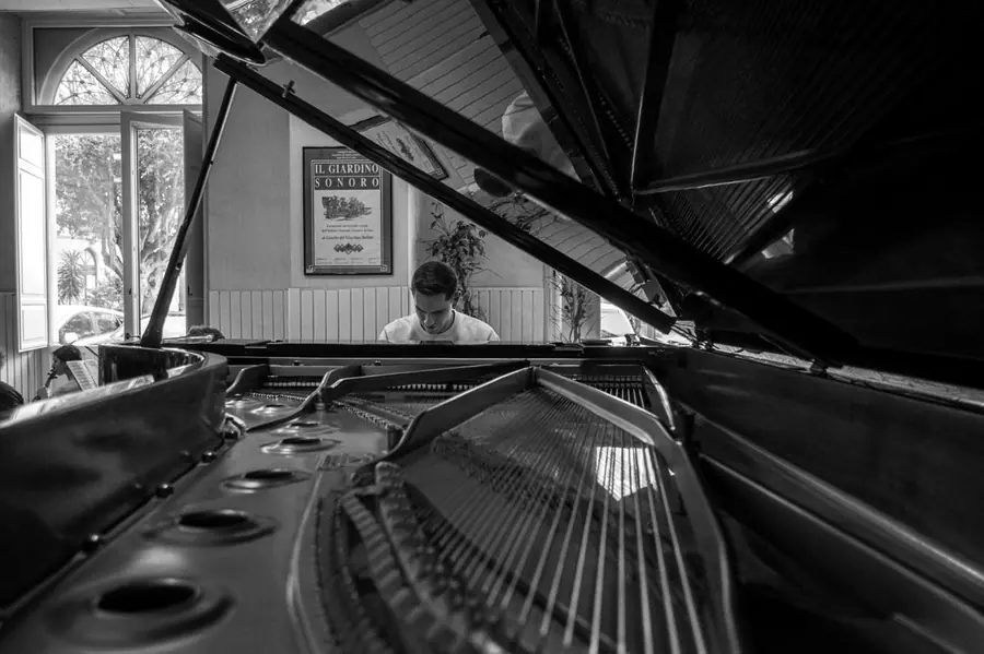 Veduta in bianco e nero dall'interno di un pianoforte a coda aperto: corde, meccanica e cerchi della tavola armonica in primo piano, oltre il leggio un giovane pianista che suona in una sala con finestra e un manifesto alla parete; non e' riconoscibile come Comis