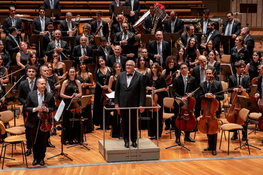 Veduta larga del palco: orchestra sinfonica in piedi con gli strumenti abbassati e il direttore in frac sulla pedana al centro, sala moderna dal palcoscenico in legno chiaro