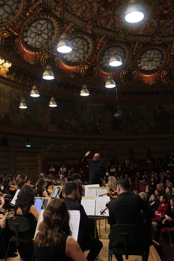Direttore sul podio con il braccio destro alzato, ripreso da dietro l'orchestra, in una sala da concerto circolare con cupola dorata e ampio fregio affrescato; pubblico in poltrone rosse tutto intorno.