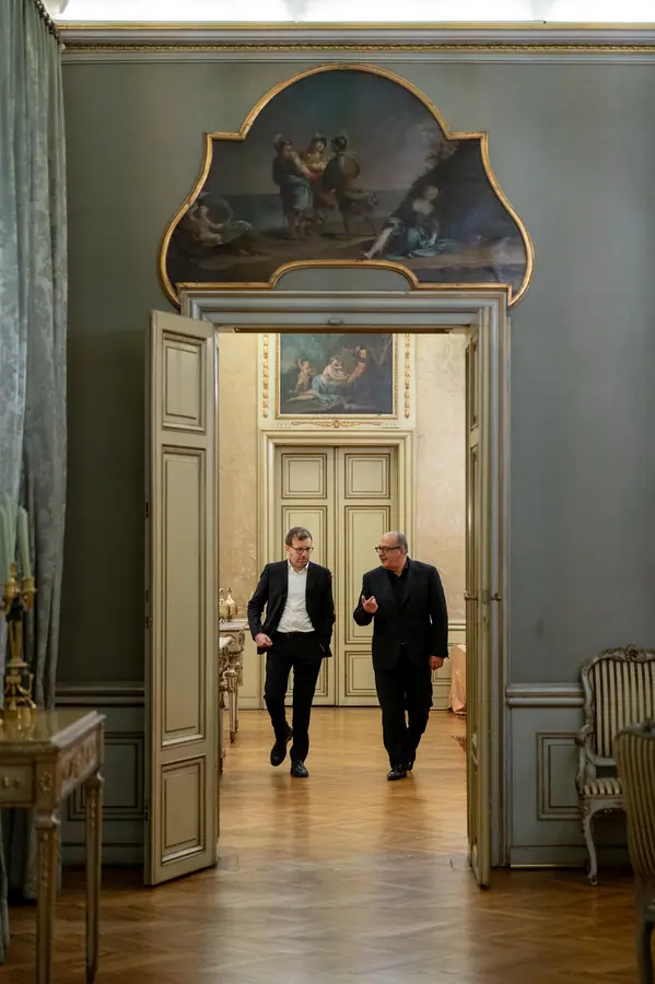 Due uomini in abito scuro camminano affiancati verso la porta aperta della sala affrescata, conversando