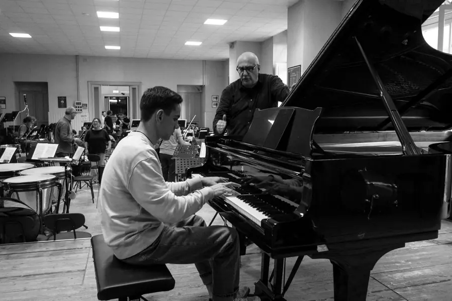 Stessa scena di prova in bianco e nero, inquadratura piu' larga: il giovane pianista di profilo alla tastiera, lui in piedi oltre il pianoforte, timpani e leggii dell'orchestra intorno